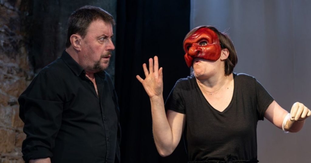 Sur scène, un homme vêtu de noir observe avec perplexité une femme qui porte un masque rouge aux traits exagérés. Les mains levées et le corps déformé dans une posture volontairement grotesque, elle semble caricaturer ou provoquer son interlocuteur. Le contraste entre son expressivité burlesque et le sérieux de l'homme crée un effet comique immédiat. Cette image met en lumière le travail de jeu au cœur de #Misanthrope, où les acteurs passent d'un personnage à l'autre et utilisent le masque, la transformation et la distanciation pour faire résonner l'humour et la satire de Molière auprès du public d'aujourd'hui.