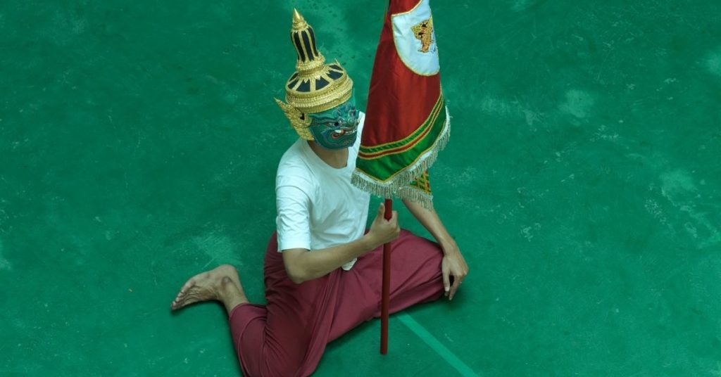 Un interprète agenouillé sur un sol vert porte un masque traditionnel thaïlandais orné d'une coiffe dorée et tient un étendard rouge. Il est vêtu d'un t-shirt blanc et d'un pantalon rouge, photographié en plongée lors d'une représentation du spectacle Miscellany of Khon