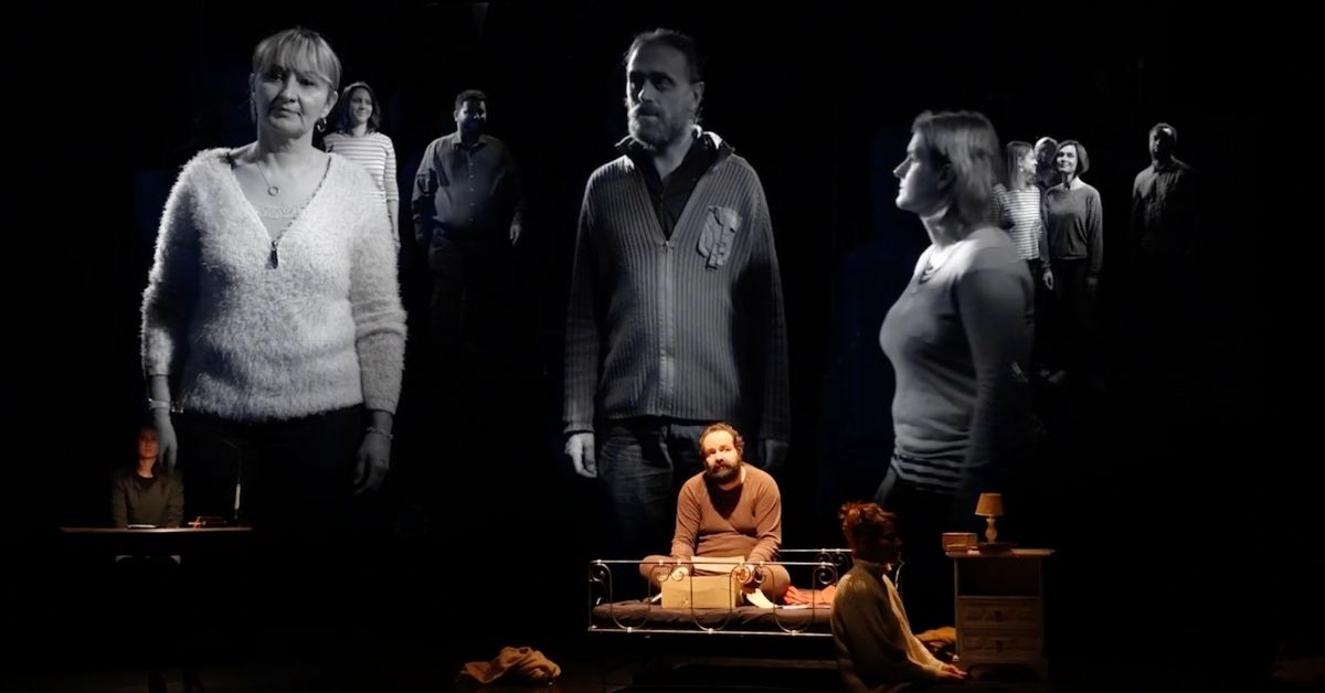 u. Au premier plan, trois personnages se tiennent autour d’un lit installé au centre de la scène, tandis que d’autres apparaissent en arrière-plan, comme des silhouettes surgissant de l’ombre. De grandes projections en noir et blanc montrent les visages et les corps des personnages, créant un effet de dédoublement entre les présences réelles et leurs images.