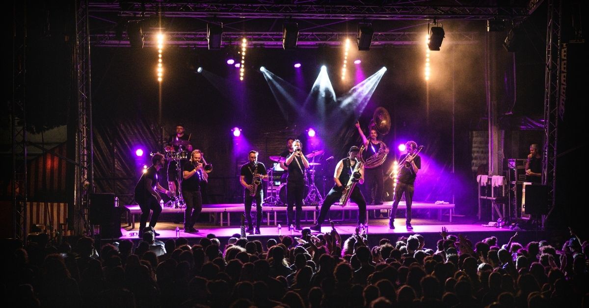 Sur scène, un ensemble de cuivres et de percussions joue devant une foule dense. Les éclairages violets, la fumée et les gestes des musiciens créent une atmosphère festive, énergique et collective.