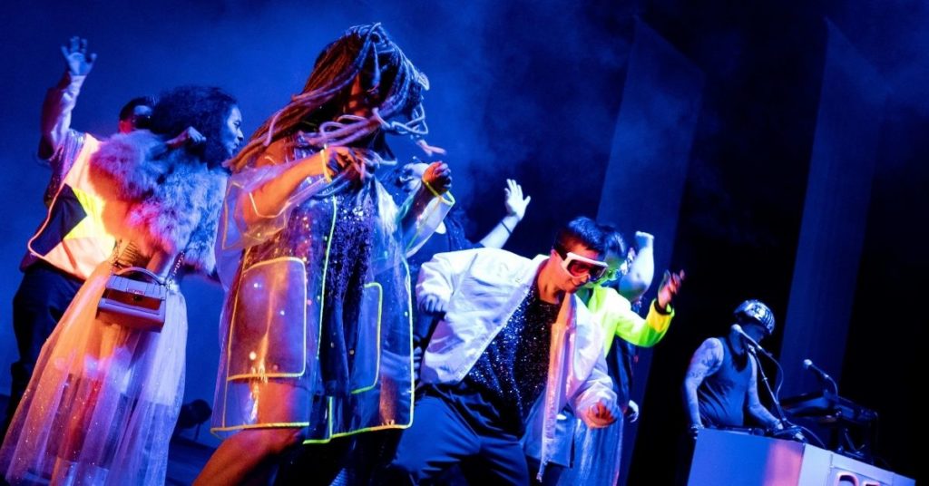 Plusieurs artistes en costumes colorés et fluorescents dansent sur scène sous un éclairage bleu, tandis qu'un musicien installé derrière une console accompagne la performance. Les tenues scintillantes et les accessoires créent une ambiance festive et électro.