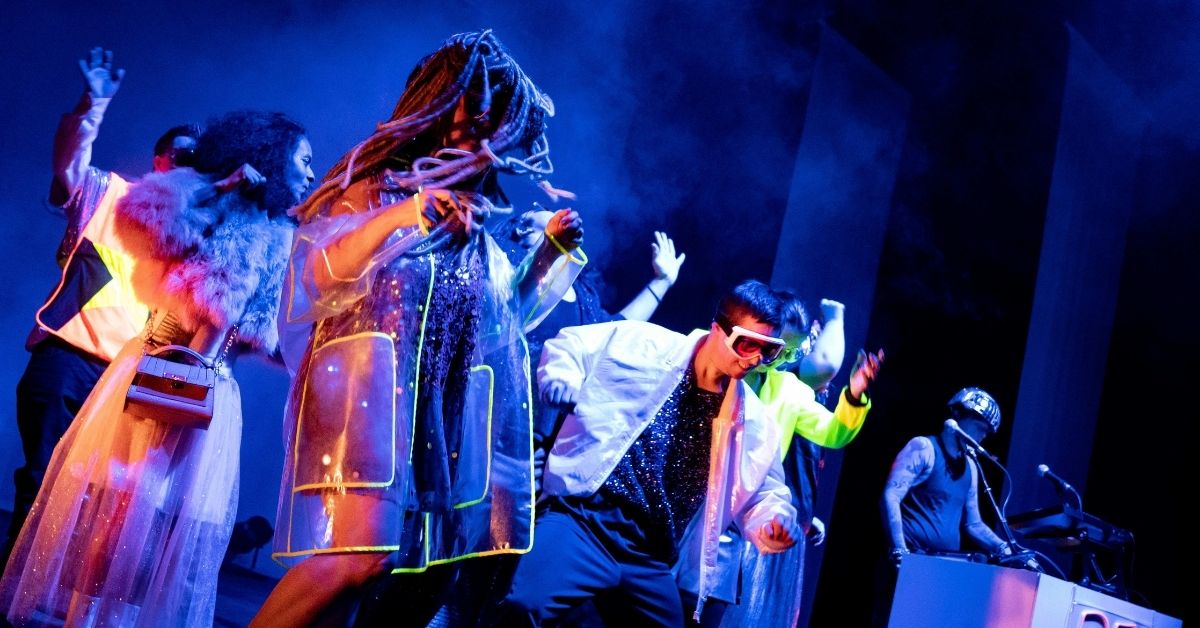 Plusieurs artistes en costumes colorés et fluorescents dansent sur scène sous un éclairage bleu, tandis qu'un musicien installé derrière une console accompagne la performance. Les tenues scintillantes et les accessoires créent une ambiance festive et électro.