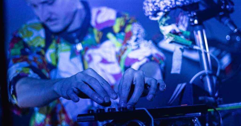 Plongé dans une lumière bleue intense, un musicien est concentré sur son instrument électronique, les mains en mouvement au-dessus des commandes. Il porte une casquette et une chemise aux motifs colorés qui contrastent avec l’atmosphère sombre et bleutée de la scène. Autour de lui, un étonnant assemblage de câbles, de machines, d’objets et d’instruments forme un univers sonore singulier.