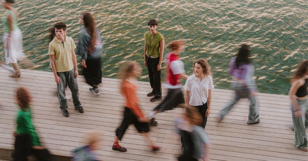 Plusieurs danseurs et danseuses se tiennent sur un ponton au bord de l'eau, tandis que d'autres traversent l'espace en mouvement flou, créant une impression de circulation et de chorégraphie.
