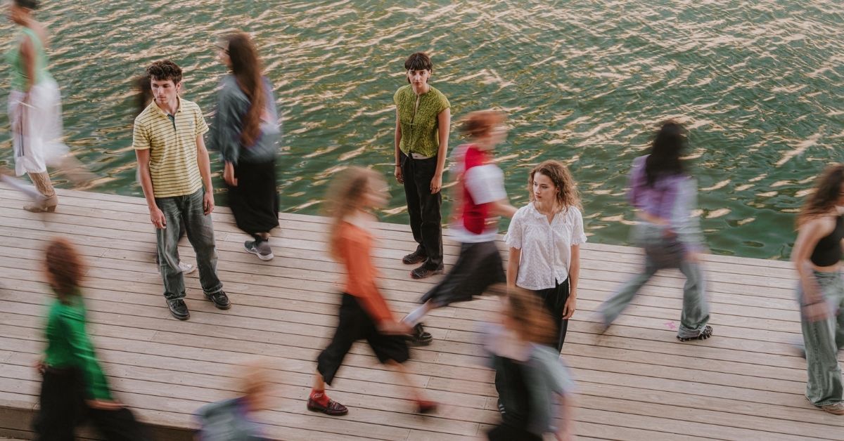 Plusieurs danseurs et danseuses se tiennent sur un ponton au bord de l'eau, tandis que d'autres traversent l'espace en mouvement flou, créant une impression de circulation et de chorégraphie.