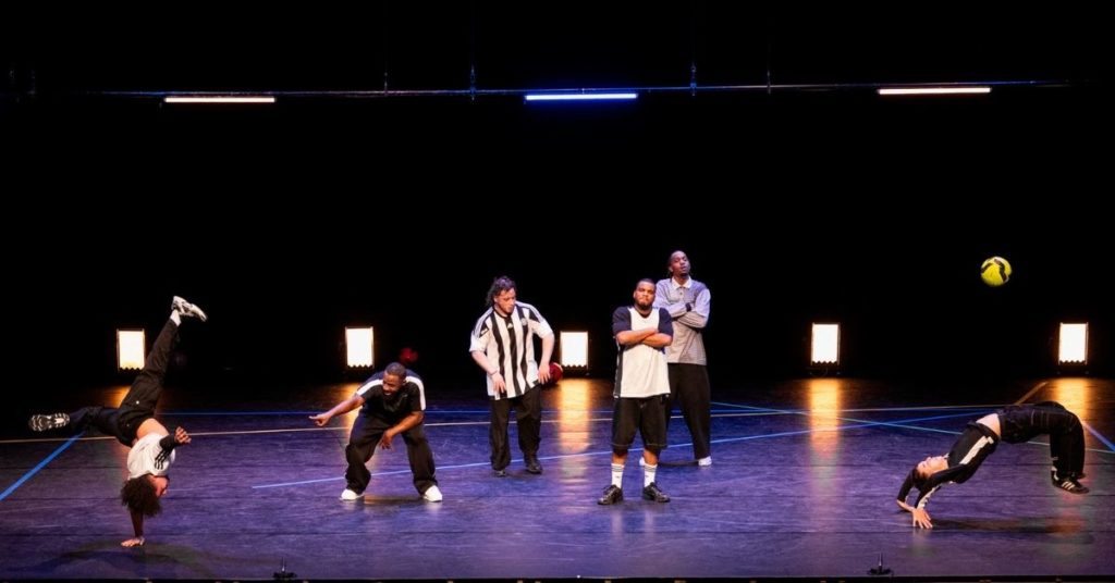 Six interprètes réalisent des figures acrobatiques et chorégraphiques autour d'un ballon de football sur une scène de théâtre, devant le public.