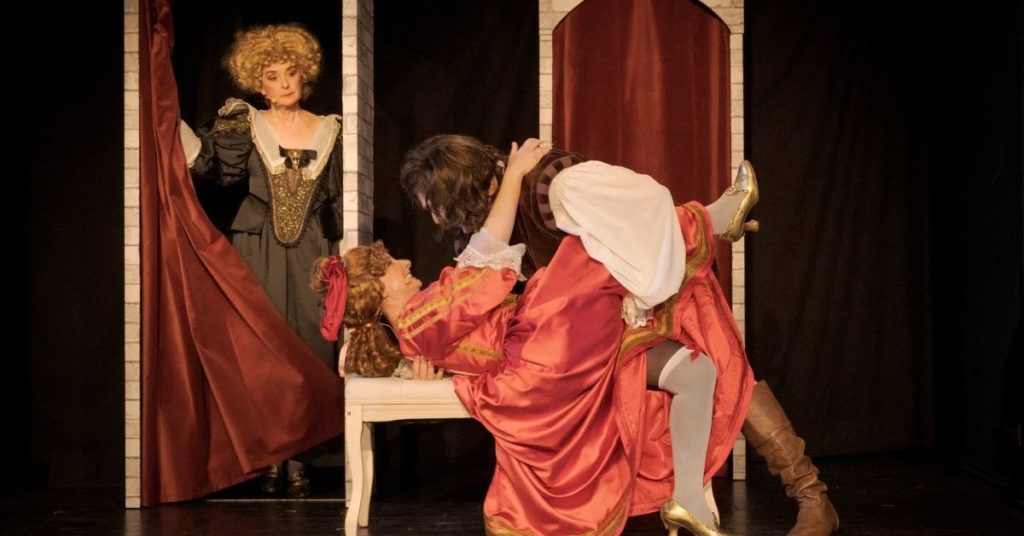 Dans un décor de palais composé d'arches et de rideaux, deux comédiens en costumes d'époque jouent une scène rapprochée au premier plan, tandis qu'un troisième personnage, vêtu d'une robe noire et d'une perruque blonde, les observe depuis une alcôve.