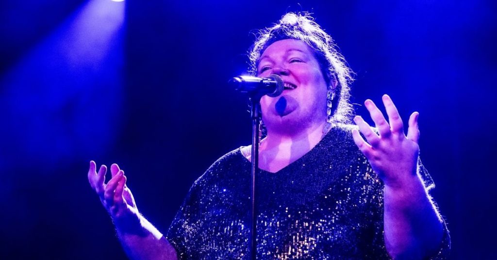 Une chanteuse interprète un morceau au micro, les mains levées, sous un éclairage de scène bleu et violet, dans une ambiance de concert intimiste.