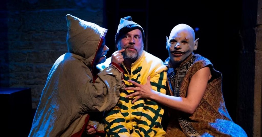 Trois comédiens en costume sont réunis sur scène sous un éclairage bleu. Au centre, un personnage vêtu d'un costume rayé jaune et vert est entouré par deux autres interprètes aux costumes et maquillages inspirés de créatures fantastiques, dans une scène expressive.