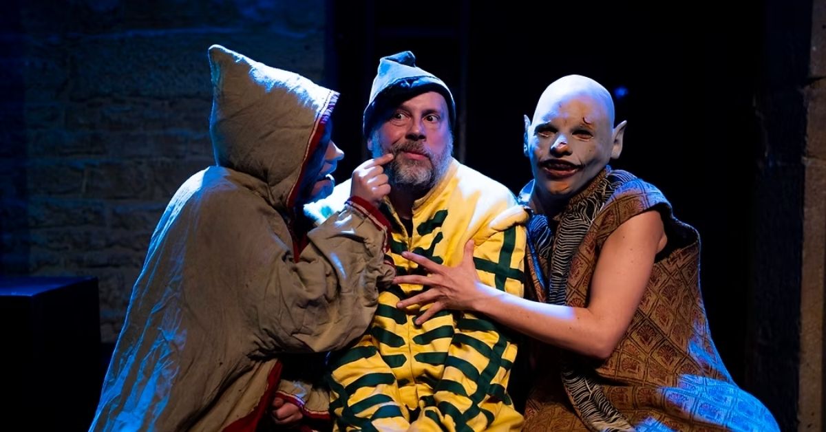 Trois comédiens en costume sont réunis sur scène sous un éclairage bleu. Au centre, un personnage vêtu d'un costume rayé jaune et vert est entouré par deux autres interprètes aux costumes et maquillages inspirés de créatures fantastiques, dans une scène expressive.