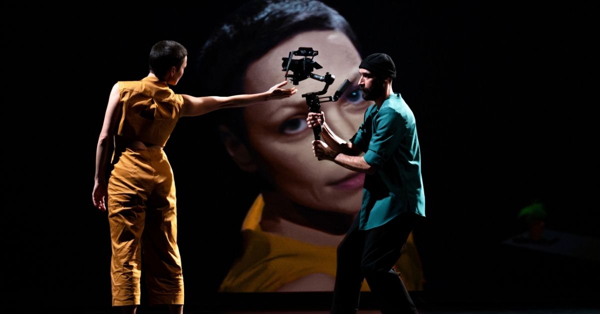 Dans une mise en scène minimaliste, une interprète tend la main vers une caméra portée par un autre interprète. Son visage, filmé en direct, est projeté en très grand format en arrière-plan, créant une atmosphère où le corps, l'image et le regard se répondent avec une troublante intimité.
