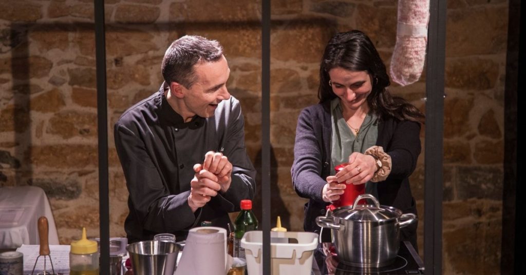 Deux interprètes préparent un repas dans une cuisine aux murs de pierre. Entre les gestes simples, les sourires échangés et la vapeur qui s'élève de la casserole, la scène fait naître une atmosphère chaleureuse où l'intime se révèle peu à peu.