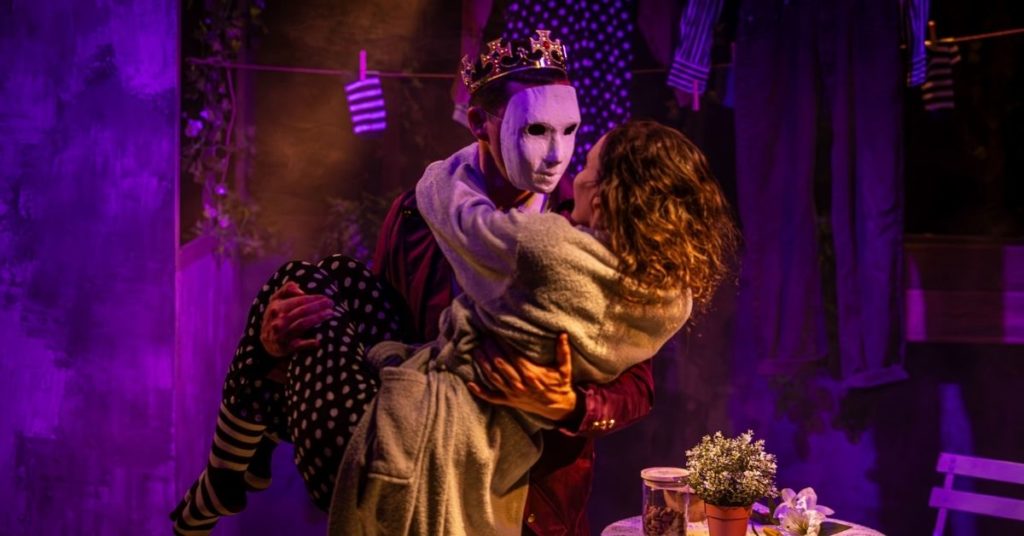 Sur une scène de théâtre baignée de lumières violettes et dorées, une personne portant un masque blanc et une couronne tient une femme dans ses bras. Les deux personnages se regardent de très près, dans une posture évoquant à la fois la tendresse et le mystère. Autour d’eux, le décor rappelle un intérieur poétique et décalé, avec du linge suspendu, une petite table recouverte d’une nappe en dentelle et quelques fleurs.