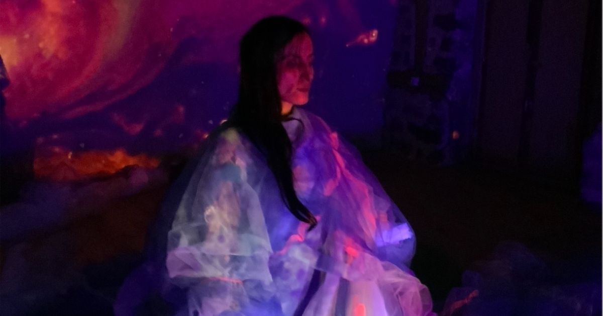 Une personne assise au centre d’un espace sombre est drapée dans de larges voiles translucides. Des projections lumineuses aux couleurs violettes, bleues et roses recouvrent à la fois le décor et les tissus, créant un univers visuel onirique.