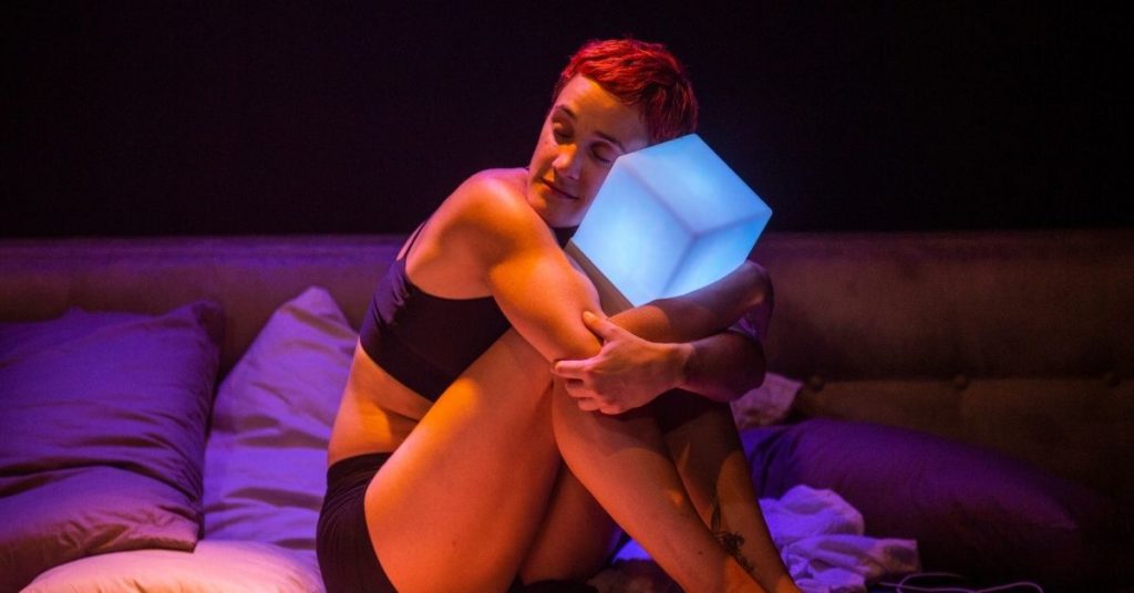 Sur un lit, une interprète enlace un cube lumineux. Une image intimiste où la douceur de la lumière contraste avec la présence énigmatique de l'objet technologique