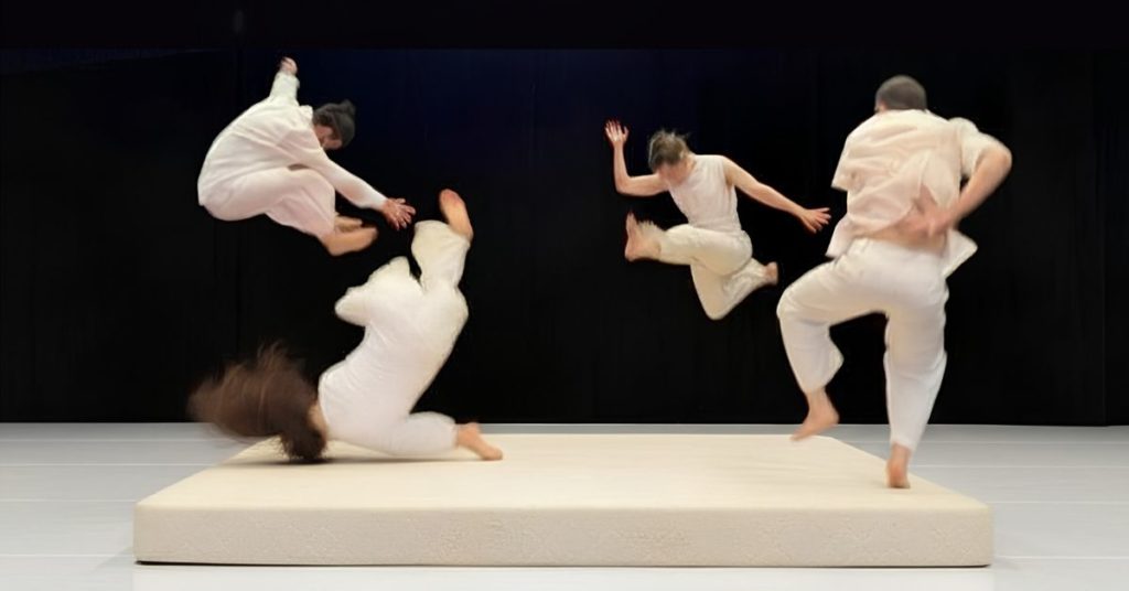 Sur un grand matelas blanc installé sur scène, quatre danseurs vêtus de blanc sont saisis dans différentes phases de saut et de chute. Leurs mouvements simultanés donnent l’impression d’un jeu collectif avec l’équilibre, la gravité et le vide.