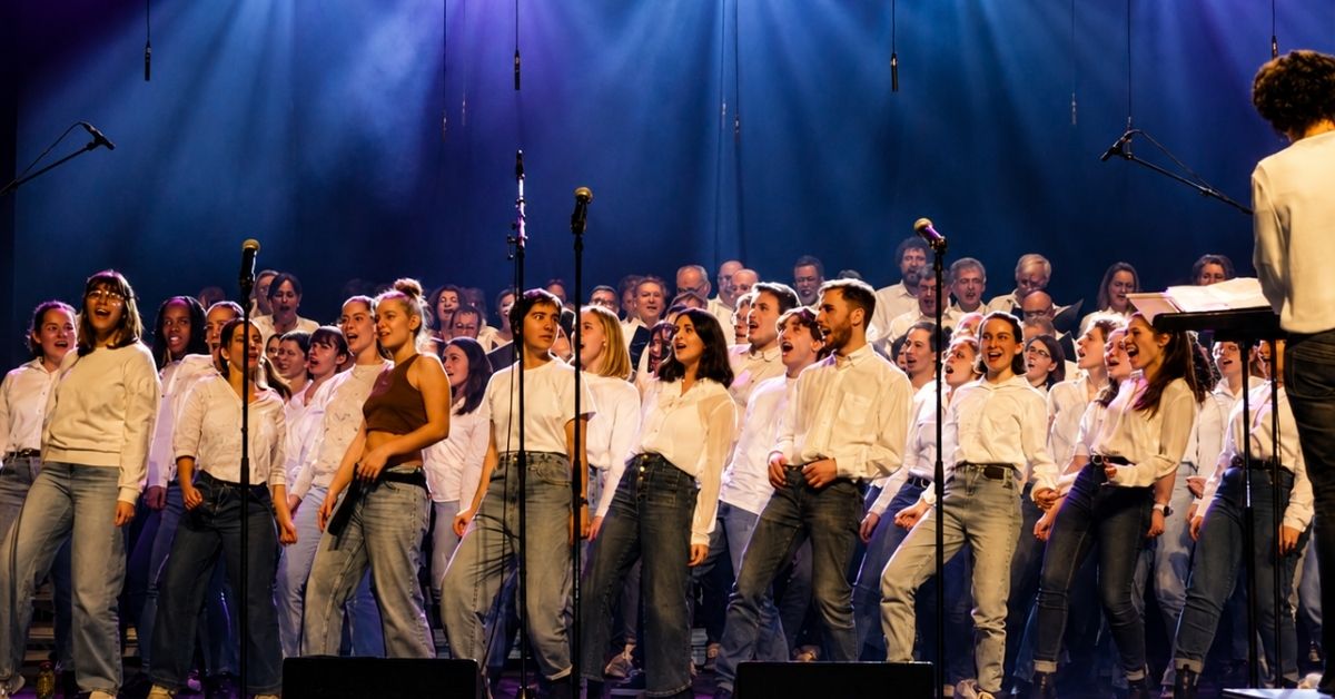 Sur une scène éclairée de bleu et de violet, un grand chœur d'une cinquantaine de jeunes chanteurs et chanteuses, vêtus principalement de blanc et de jeans, interprète un morceau devant plusieurs micros. À droite, une personne dirige le groupe depuis un pupitre.