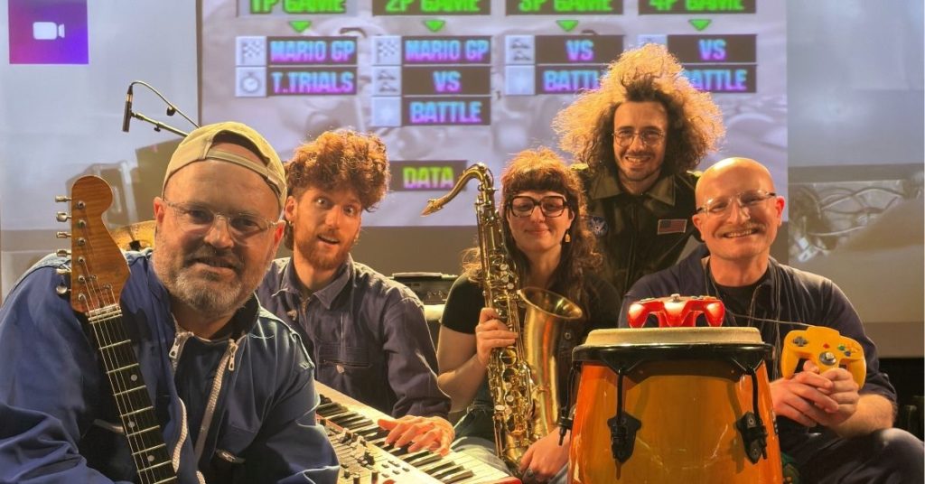 Cinq musiciennes et musiciens posent sur une scène de concert entourés de leurs instruments : guitare électrique, clavier, saxophone et percussions. Derrière eux, un écran géant affiche le menu du jeu vidéo Mario Kart 64. Les artistes sourient à l’objectif dans une ambiance conviviale et décontractée, mêlant univers du jeu vidéo et performance musicale.