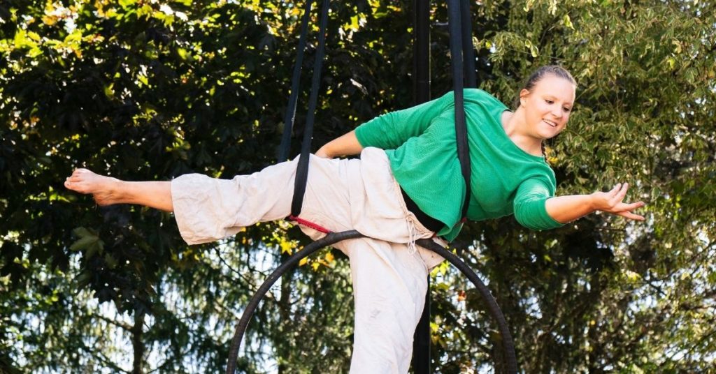 Une artiste de cirque évolue dans un cerceau aérien suspendu entre les arbres. Souriante, elle est allongée en équilibre, un bras tendu vers l'avant, vêtue d'un haut vert et d'un pantalon clair.