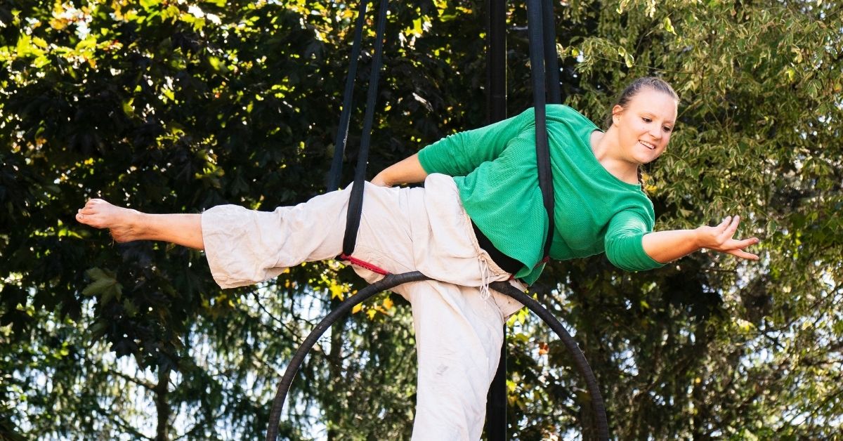 Une artiste de cirque évolue dans un cerceau aérien suspendu entre les arbres. Souriante, elle est allongée en équilibre, un bras tendu vers l'avant, vêtue d'un haut vert et d'un pantalon clair.