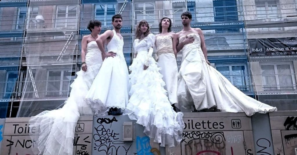 Cinq interprètes posent en robes de mariée devant une façade urbaine en rénovation recouverte d’échafaudages. Le contraste entre les codes traditionnels du mariage et ce décor brut évoque le regard critique porté par le spectacle sur les modèles amoureux, conjugaux et familiaux hérités, au cœur de l’adaptation de la bande dessinée de Liv Strömquist.