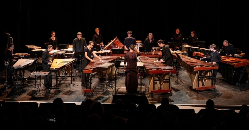 Une quinzaine d’interprètes occupent la scène entourés de marimbas, vibraphones, timbales et autres instruments de percussion. L’organisation de l’ensemble, centrée autour de la direction d’orchestre, met en valeur l’ampleur de cette formation singulière