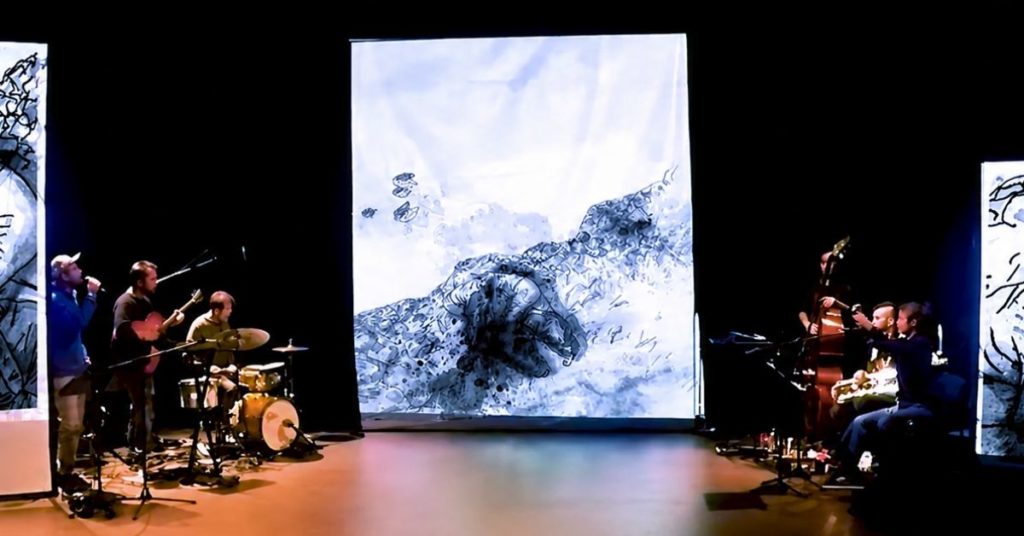 Une scène de spectacle plongée dans la pénombre, où plusieurs musiciens se produisent de part et d'autre de l'espace scénique. Trois grands écrans verticaux projettent des illustrations en noir et blanc, réalisées dans un style mêlant encre et aquarelle.