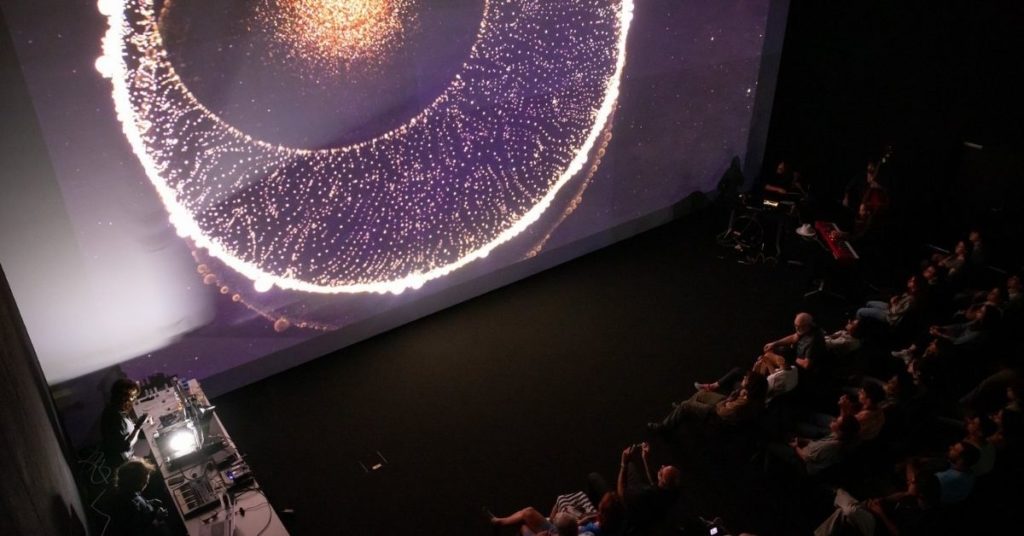 Face à une projection monumentale de formes lumineuses rappelant à la fois l’infiniment petit et l’infiniment grand, le public assiste à une performance où images scientifiques et musique live se rencontrent dans une atmosphère contemplative et fascinante.