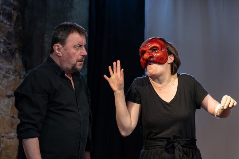 Sur scène, un homme vêtu de noir observe avec perplexité une femme qui porte un masque rouge aux traits exagérés. Les mains levées et le corps déformé dans une posture volontairement grotesque, elle semble caricaturer ou provoquer son interlocuteur. Le contraste entre son expressivité burlesque et le sérieux de l'homme crée un effet comique immédiat. Cette image met en lumière le travail de jeu au cœur de #Misanthrope, où les acteurs passent d'un personnage à l'autre et utilisent le masque, la transformation et la distanciation pour faire résonner l'humour et la satire de Molière auprès du public d'aujourd'hui.
