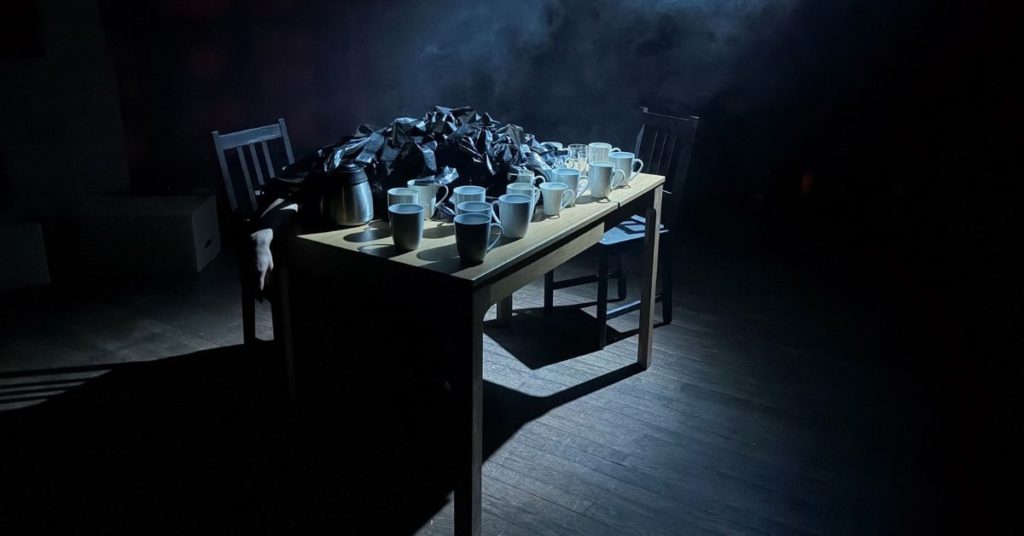 Dans une salle sombre et brumeuse, une table est positionnée au centre1. Sur la table, une dizaine de tasse et une personne ensevelie sous du plastique.