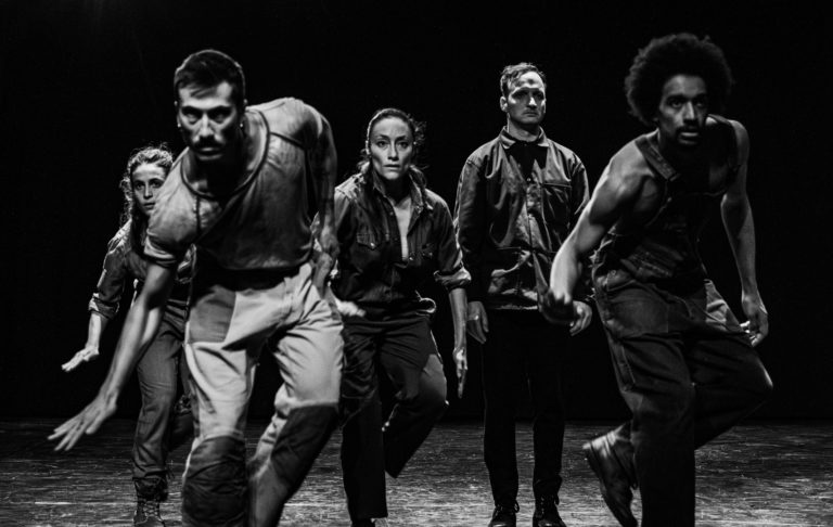 Cinq interprètes occupent la scène dans une formation resserrée, avançant d’un même élan vers le public. Leurs postures engagées, leurs regards intenses et leurs mouvements synchronisés traduisent l’énergie collective propre au stepping, où le corps devient instrument de percussion. La photographie en noir et blanc met en valeur la force physique, la précision rythmique et les dynamiques de groupe qui sont au cœur du spectacle Isicathulo.