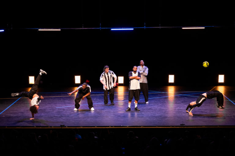 Six interprètes réalisent des figures acrobatiques et chorégraphiques autour d'un ballon de football sur une scène de théâtre, devant le public.