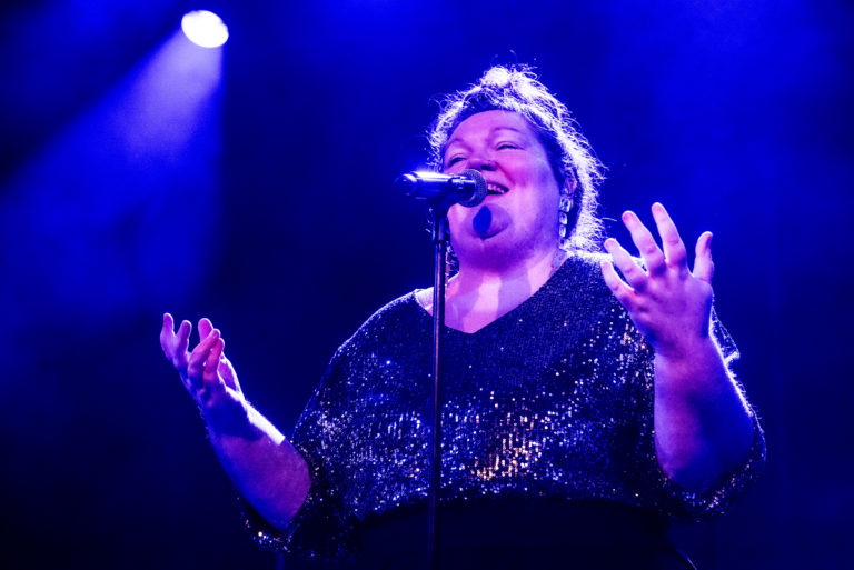 Une chanteuse interprète un morceau au micro, les mains levées, sous un éclairage de scène bleu et violet, dans une ambiance de concert intimiste.
