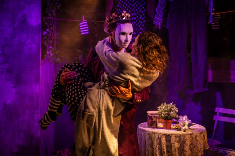 Sur une scène de théâtre baignée de lumières violettes et dorées, une personne portant un masque blanc et une couronne tient une femme dans ses bras. Les deux personnages se regardent de très près, dans une posture évoquant à la fois la tendresse et le mystère. Autour d’eux, le décor rappelle un intérieur poétique et décalé, avec du linge suspendu, une petite table recouverte d’une nappe en dentelle et quelques fleurs.