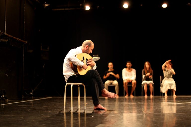Sur une scène de théâtre plongée dans la pénombre, une personne assise pieds nus sur une chaise joue du oud, un instrument à cordes oriental, sous un éclairage chaleureux. À l’arrière-plan, quatre autres interprètes, également pieds nus, sont assis et légèrement flous. L’un d’eux effectue un geste avec les bras. L’espace scénique est sobre et épuré, mettant en valeur la présence des corps et du musicien.