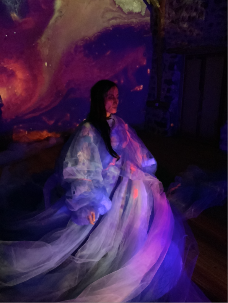 Une personne assise au centre d’un espace sombre est drapée dans de larges voiles translucides. Des projections lumineuses aux couleurs violettes, bleues et roses recouvrent à la fois le décor et les tissus, créant un univers visuel onirique.