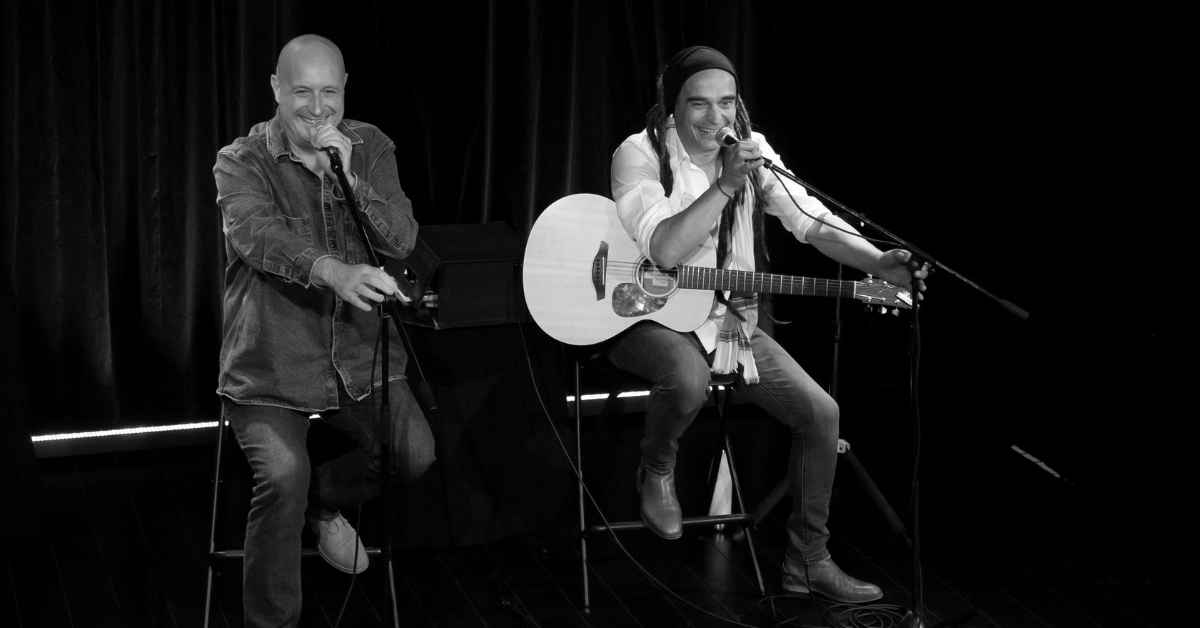 Sur une scène plongée dans une lumière sobre, deux artistes sont assis côte à côte sur de hauts tabourets. Ils chantent dans des microphones et semblent partager un moment complice avec le public. L’un tient une guitare acoustique blanche, tandis que l’autre accompagne la performance au chant. Leurs sourires et leurs regards donnent à la scène une atmosphère chaleureuse, conviviale et légère