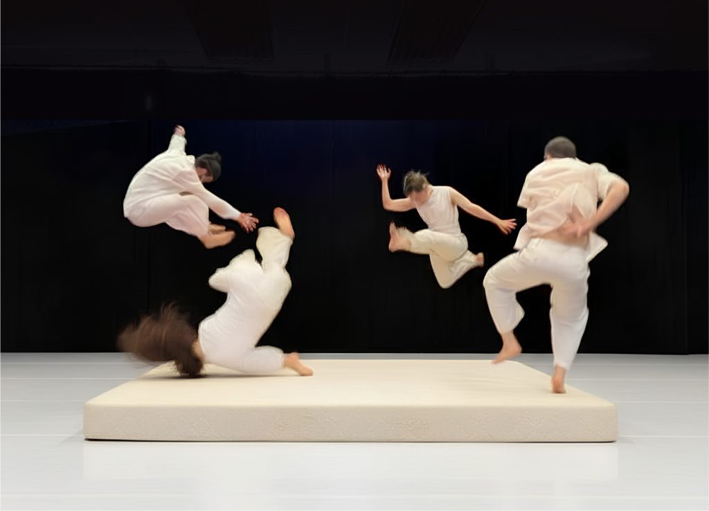 Sur un grand matelas blanc installé sur scène, quatre danseurs vêtus de blanc sont saisis dans différentes phases de saut et de chute. Leurs mouvements simultanés donnent l’impression d’un jeu collectif avec l’équilibre, la gravité et le vide.