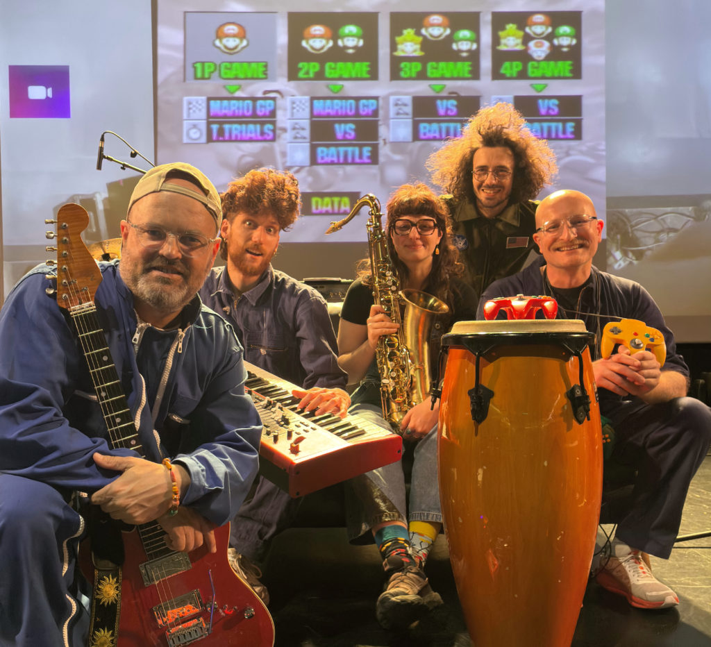 Cinq musiciennes et musiciens posent sur une scène de concert entourés de leurs instruments : guitare électrique, clavier, saxophone et percussions. Derrière eux, un écran géant affiche le menu du jeu vidéo Mario Kart 64. Les artistes sourient à l’objectif dans une ambiance conviviale et décontractée, mêlant univers du jeu vidéo et performance musicale.