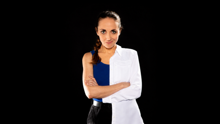 Portrait d'une comédienne debout sur fond noir, les bras croisés, vêtue d'un débardeur bleu et d'une blouse blanche portée sur une seule moitié du corps, regardant l'objectif avec un léger sourire.