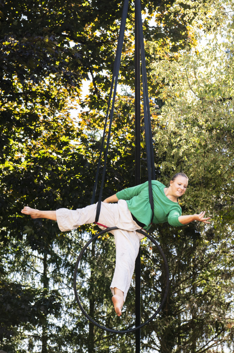 Une artiste de cirque évolue dans un cerceau aérien suspendu entre les arbres. Souriante, elle est allongée en équilibre, un bras tendu vers l'avant, vêtue d'un haut vert et d'un pantalon clair.