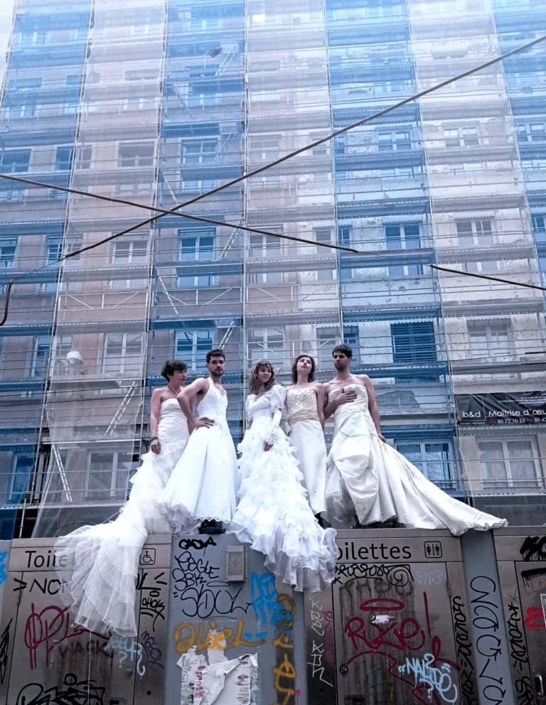 Cinq interprètes posent en robes de mariée devant une façade urbaine en rénovation recouverte d’échafaudages. Le contraste entre les codes traditionnels du mariage et ce décor brut évoque le regard critique porté par le spectacle sur les modèles amoureux, conjugaux et familiaux hérités, au cœur de l’adaptation de la bande dessinée de Liv Strömquist.