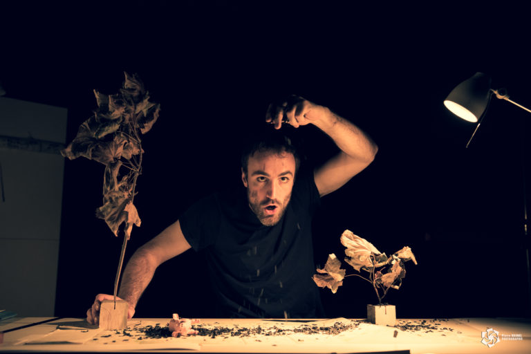 Un homme, éclairé par une lampe de bureau, est penché au-dessus d'une table recouverte de terre. Il tient un objet suspendu au-dessus d'un décor miniature composé de deux petits arbres aux feuilles sèches, dans une scène de théâtre sur fond noir.