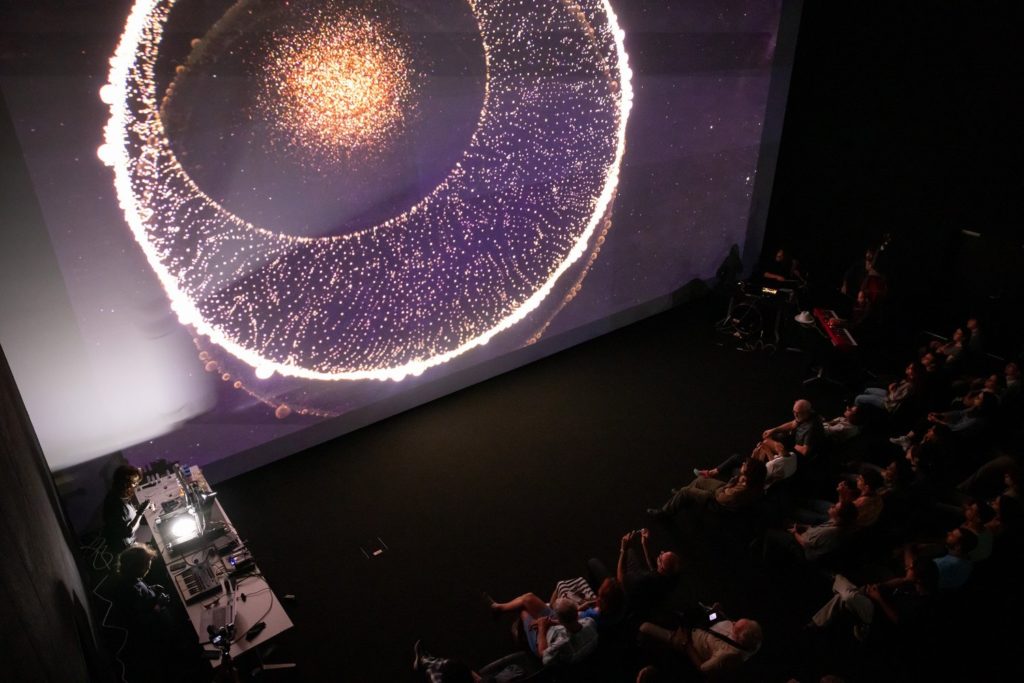 Face à une projection monumentale de formes lumineuses rappelant à la fois l’infiniment petit et l’infiniment grand, le public assiste à une performance où images scientifiques et musique live se rencontrent dans une atmosphère contemplative et fascinante.