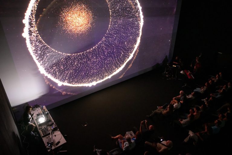 Face à une projection monumentale de formes lumineuses rappelant à la fois l’infiniment petit et l’infiniment grand, le public assiste à une performance où images scientifiques et musique live se rencontrent dans une atmosphère contemplative et fascinante.