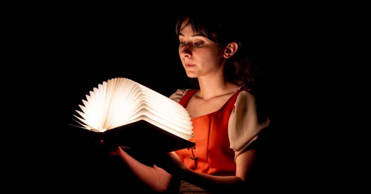 Une jeune femme est assise dans l’obscurité et tient devant elle un grand livre ouvert dont les pages blanches diffusent une lumière douce sur son visage et ses bras. Elle porte une robe rouge orangé sur un chemisier clair à manches bouffantes. Son regard est dirigé vers le livre, tandis que le fond entièrement noir crée une atmosphère intime, mystérieuse et théâtrale. Le contraste entre la lumière du livre et l’obscurité environnante met en valeur la lectrice et l’objet qu’elle tient.